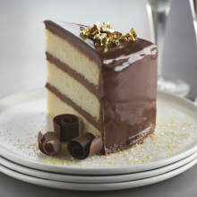 A slice of layered vanilla cake with glossy chocolate icing, topped with gold flakes and chocolate curls, sits on a white plate with gold sprinkles. Two glasses in the background suggest a celebratory setting.