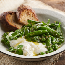 A rustic bowl with creamy burrata cheese, fresh green peas, snap peas, herbs, and a drizzle of olive oil, served with two slices of toasted bread on the side. The bowl rests on a wooden table.