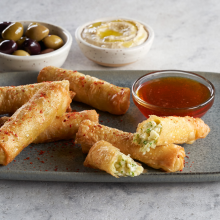 Six crispy rolls, some broken open to reveal a vegetable filling, on a gray plate with a bowl of red dipping sauce. In the background, bowls of mixed olives and hummus are visible.