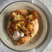 Roasted cauliflower florets served on a creamy orange sauce, topped with a dollop of white sauce and sprinkled with chopped herbs, on a rustic beige plate over a light napkin and wooden surface.