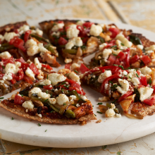 A round vegetable pizza topped with diced tomatoes, green bell peppers, onions, and crumbled cheese, sliced into six pieces and served on a white marble platter.