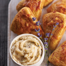 Golden scones on a white wooden board next to a small bowl of herb butter, garnished with sprigs of fresh lavender.