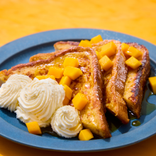 Three slices of French toast topped with diced mango and syrup, served with two swirls of whipped cream on a blue plate, set against a bright yellow background.