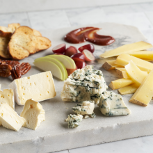 A marble board with assorted cheeses, sliced apples, grapes, pecans, sliced bread crisps, and fruit preserves arranged neatly.