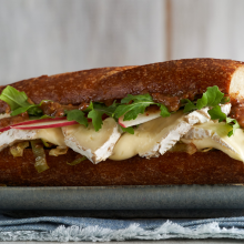 A close-up of a sandwich on a baguette with melted brie cheese, sliced apples, leafy greens, caramelized onions, and a spread, served on a plate over a blue cloth.