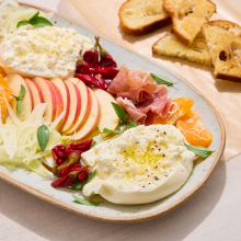 A platter with sliced apples, tangerine segments, fennel, prosciutto, spicy peppers, fresh burrata cheese drizzled with olive oil, and herbs, served with toasted bread slices on the side.