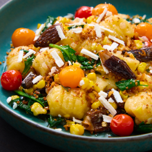 A blue plate filled with gnocchi, cherry tomatoes, spinach, corn, morel mushrooms, and crumbs, topped with grated cheese. The colorful ingredients are attractively arranged, creating a vibrant, appetizing dish.