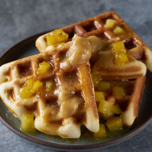 Two golden brown waffles topped with diced caramelized pineapple and syrup are served on a dark round plate, placed on a gray textured surface.