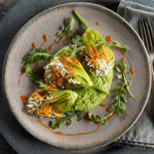 Three stuffed zucchini blossoms served on a light green sauce, garnished with arugula and a drizzle of red sauce, placed on a gray plate with a fork and napkin on the side.