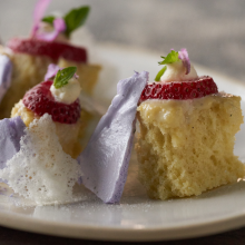 A close-up of a plated dessert featuring sponge cake pieces topped with whipped cream, strawberry slices, small green leaves, and purple meringue shards on a white plate.