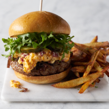 A cheeseburger topped with arugula, pickles, and a creamy spread, served on a bun with a side of golden French fries on a marble surface.
