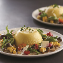 A plated dish featuring a yellow custard dome surrounded by arugula, cherry tomatoes, grilled corn, mushrooms, parmesan shavings, and microgreens. A second, similar plate is blurred in the background.