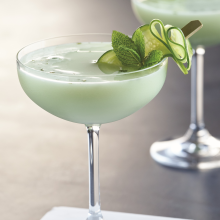 A pale green cocktail served in a coupe glass, garnished with cucumber slices and a sprig of mint. Another similar drink is blurred in the background. The scene is set on a light surface.