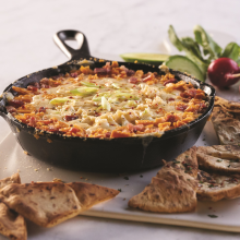 A skillet filled with baked cheesy dip topped with chopped green onions, surrounded by toasted pita bread slices, fresh radishes, lime wedges, and greens on a white serving board.