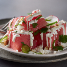 A plate of fresh watermelon cubes topped with white cheese, cream drizzle, sliced radishes, cilantro, and nuts, served with a fork on a ceramic plate.
