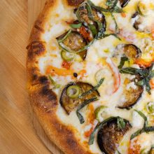Close-up of a pizza topped with melted cheese, slices of eggplant, green onions, red and yellow bell peppers, and fresh basil, on a wooden surface.