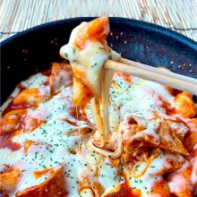 A pan filled with tteokbokki, covered in melted cheese, with a chopstick lifting a piece, stretching gooey cheese above the dish.