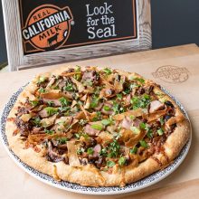 A pizza topped with cheese, green herbs, and sliced toppings sits on a plate. In the background, a sign reads "Real California Milk" and "Look for the Seal." The pizza is on a wooden board.