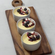 Creamy yellow custard dessert bowls topped with fresh berries and berry sauce on a wooden board.