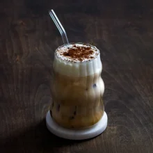 A classic Einspanner latte drink with layered espresso and cream, topped with cocoa powder dusting, served in a clear glass with straw.