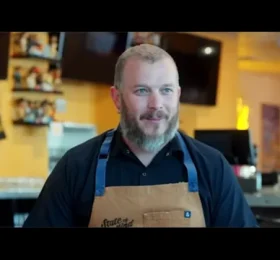 Male food service worker wearing branded apron in commercial kitchen setting, representing professional foodservice and wholesale food industry operations.