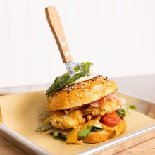 Gourmet burger with crispy chicken, vibrant toppings, and toasted sesame seed bun on a tray.