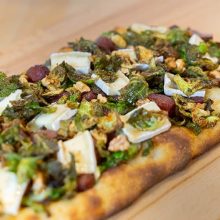 A rectangular flatbread topped with melted brie cheese, Brussels sprouts, walnuts, and pieces of sausage, placed on a wooden surface.