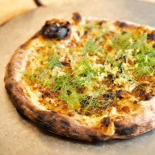 A rustic, wood-fired pizza topped with caramelized onions, fresh dill, lemon zest, and a charred garlic bulb, served on a metal tray.