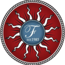 A circular emblem with a red background, stylized black and white sun rays, and a blue center featuring a white script "F" and the text "EST. 1985," bordered by a rope-like pattern, inspired by California Dairies heritage.
