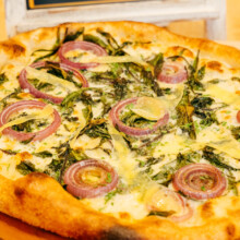 Artisan pizza topped with sautéed greens, cheese, and red onion on a golden-brown crust.