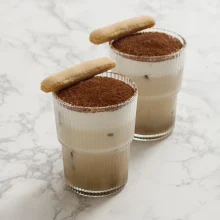 Two tiramisu latte desserts in commercial glassware topped with cocoa powder and ladyfinger cookies, ideal for food service and wholesale distribution.