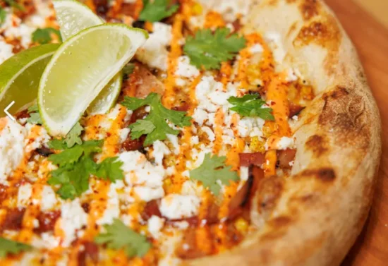 Grilled corn elote topped with crumbled queso fresco, chili powder, cilantro and lime wedge on charred tortilla.