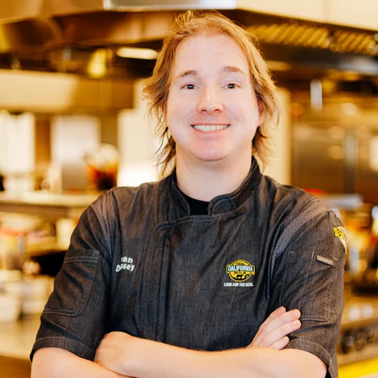 Professional headshot of a smiling food service industry ambassador in branded black chef coat in commercial kitchen setting.