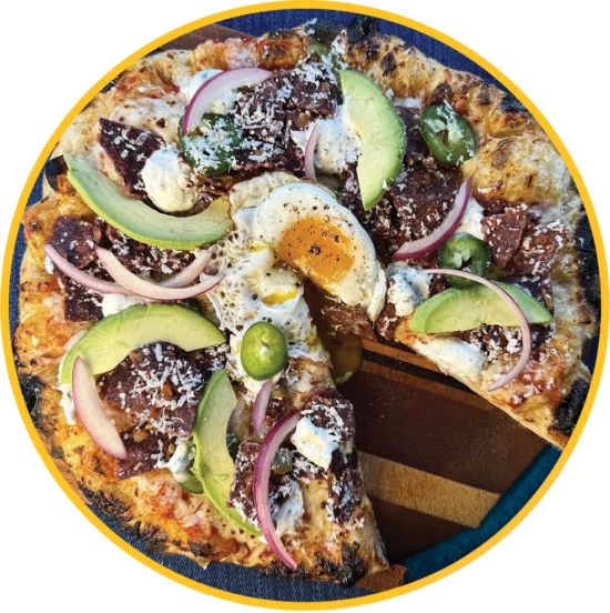 Artisanal breakfast pizza topped with runny eggs, avocado slices, red onion, jalapeños, and queso fresco on charred crust.