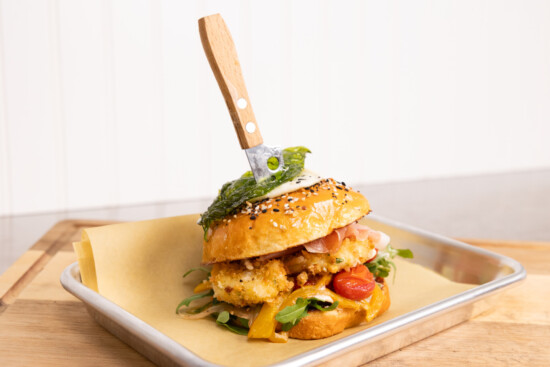 Gourmet burger with crispy chicken, vibrant toppings, and toasted sesame seed bun on a tray.