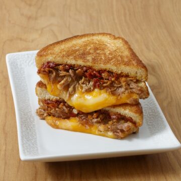 Golden-brown grilled cheese sandwich filled with shredded carnitas, melted Colby Jack cheese, and caramelized onions, plated on white porcelain for foodservice.