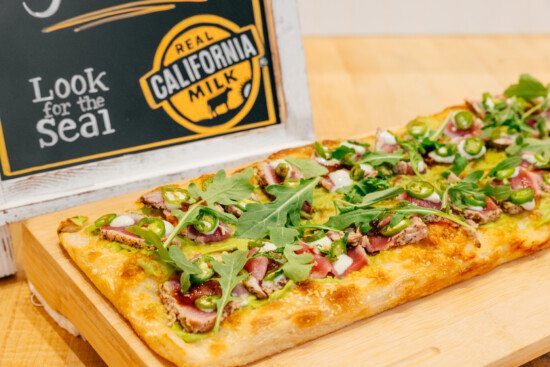 Delicious rectangular pizza topped with avocado, seared meat, arugula, and jalapeños.