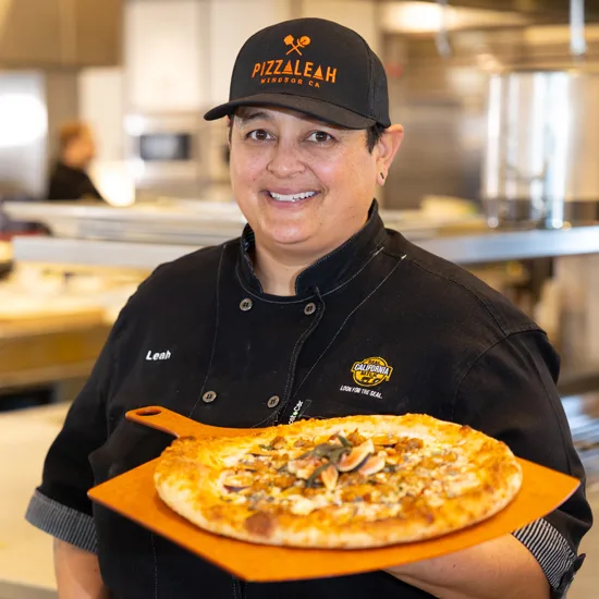 PizzaLeah restaurant staff member proudly displaying a freshly prepared pizza on an orange plate in commercial kitchen setting.