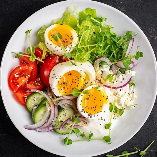 A vibrant cottage cheese salad bowl featuring three sunny-side eggs, fresh greens, sliced cucumbers, red tomatoes, and microgreens on a white plate.