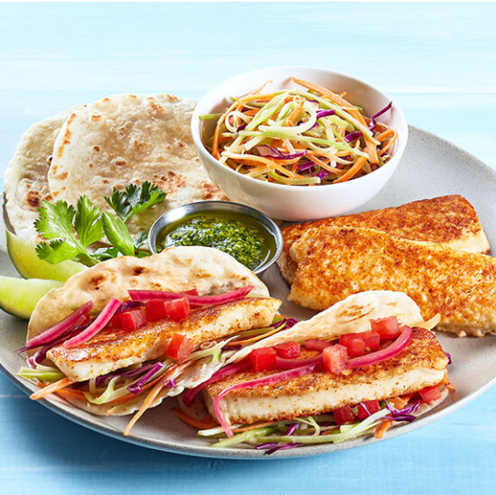 A plate with two fish tacos topped with shredded cabbage, onions, and tomatoes, grilled fish fillets, a bowl of coleslaw, flatbreads, a lime wedge, cilantro, and a small container of green sauce.