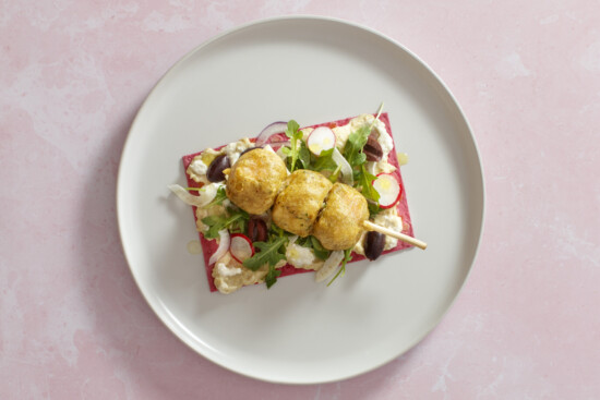 Vibrant gourmet dish with arugula, radishes, olives, and golden protein skewers on a colorful base.